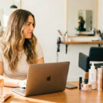 A woman working on a laptop at a desk in a modern salon, surrounded by beauty products and a camera, illustrating non-service beauty careers like content creation or salon management that don't require a license.