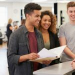 A diverse group of young adults holding enrollment folders while talking to an admissions representative at a modern beauty school front desk with a salon floor in the background.