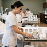 A student in a white smock organizes skincare tools and bowls on a rolling cart beside a reclining treatment bed.
