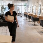 First time salon owner holding a blank permit folder while walking through a nearly finished salon with floor plans, covered mirrors, styling stations, and shampoo bowls.