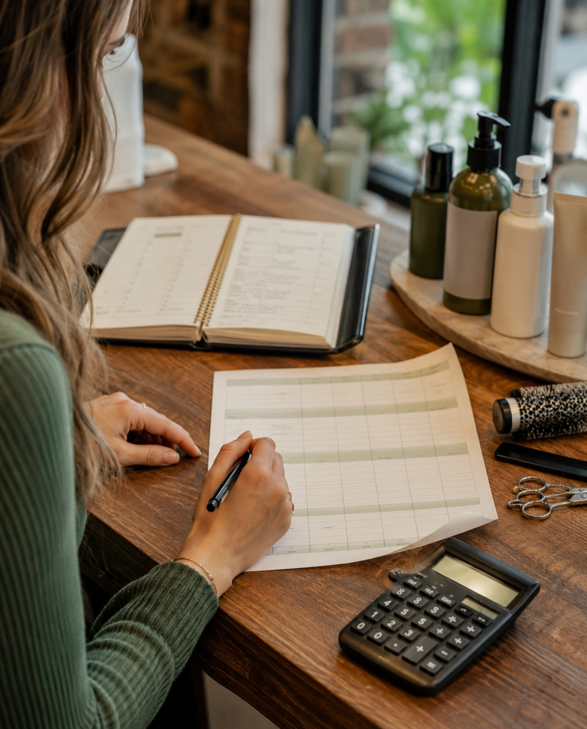 New stylist reviewing an unreadable earnings worksheet with a calculator, appointment book, blank product bottles, and salon tools.