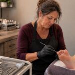 Experienced nail technician performs a careful waterless pedicure for an older client beside a closed laptop used for advanced professional training.