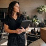 An experienced cosmetologist holds a license portfolio while packing a salon station, rolling tool case, moving box, and road map.