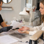 Nail technician reviews a client intake form while examining bare natural hands at a manicure station with a policy document and clean equipment.