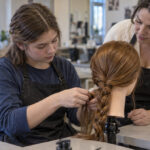 High school student practicing a three strand braid on a mannequin while an adult cosmetology instructor provides hands on guidance in a vocational classroom.
